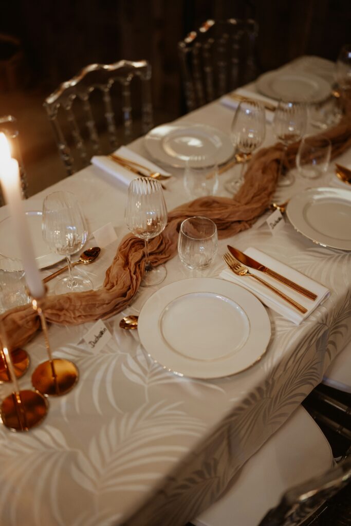A refined holiday dinner table set with gold flatware, glass stemware, white plates, and a soft taupe runner on a patterned ivory tablecloth, creating a warm and elegant ambiance.