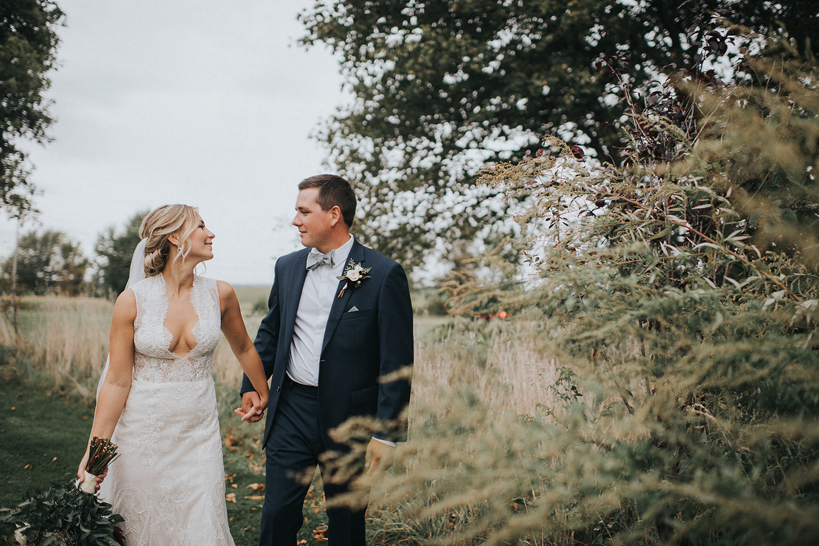 Newlywed couple holding hands and walking through a grassy field during golden hour portraits at The Wells Estate wedding venue.