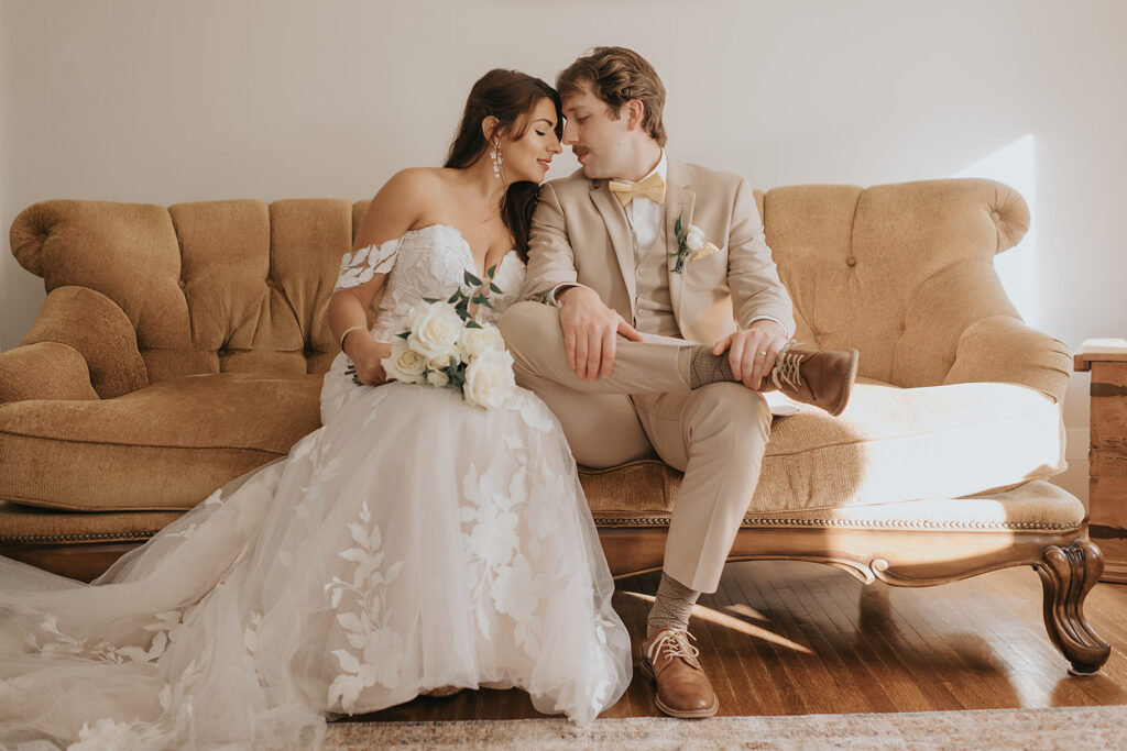 Bride and groom sitting close together on a tan vintage couch indoors at The Wells Estate with soft natural light.