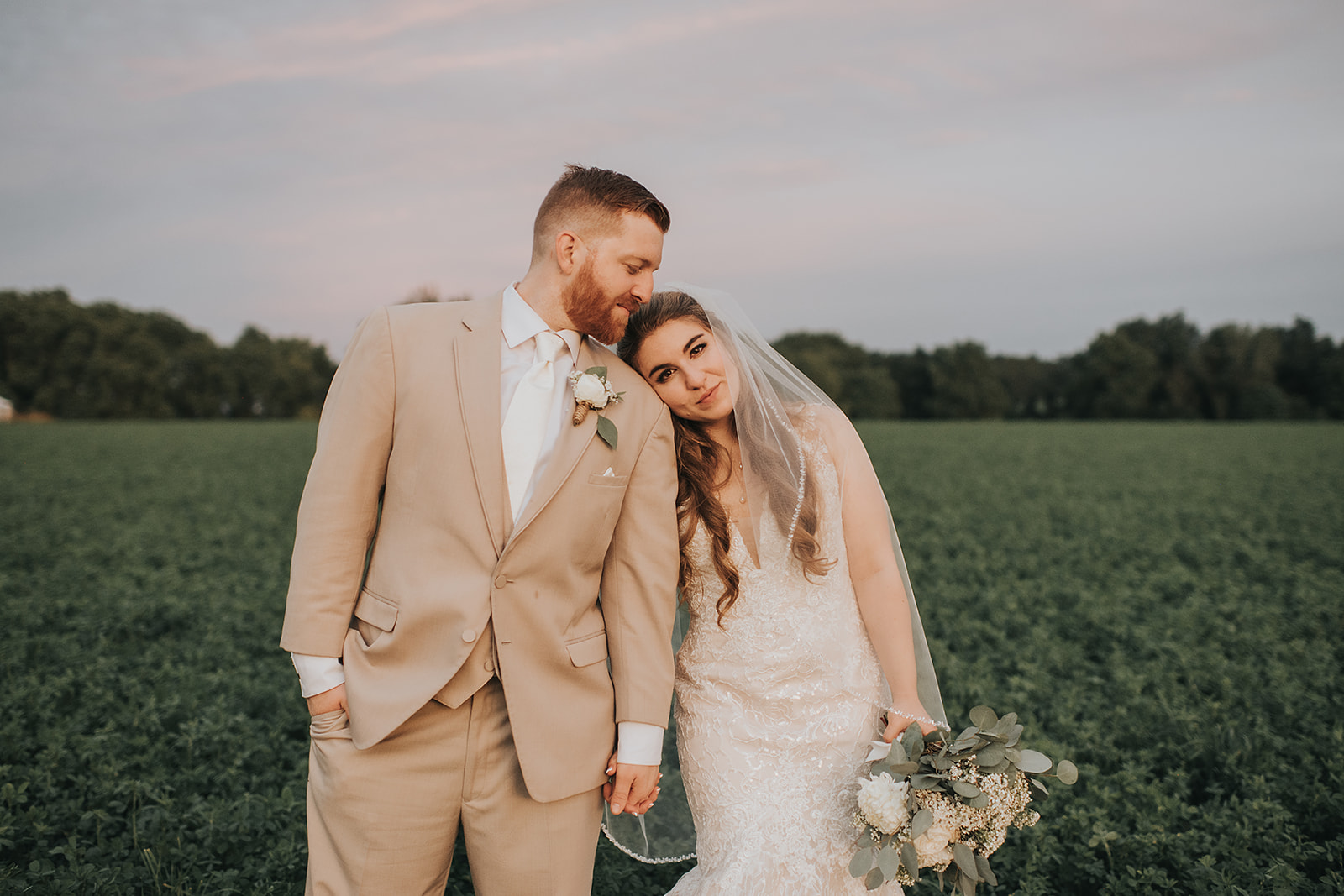 Bride and groom standing hand in hand in a lush green field at sunset, the bride resting her head on the groom’s shoulder while holding a eucalyptus and white floral bouquet at The Wells Estate wedding venue.
