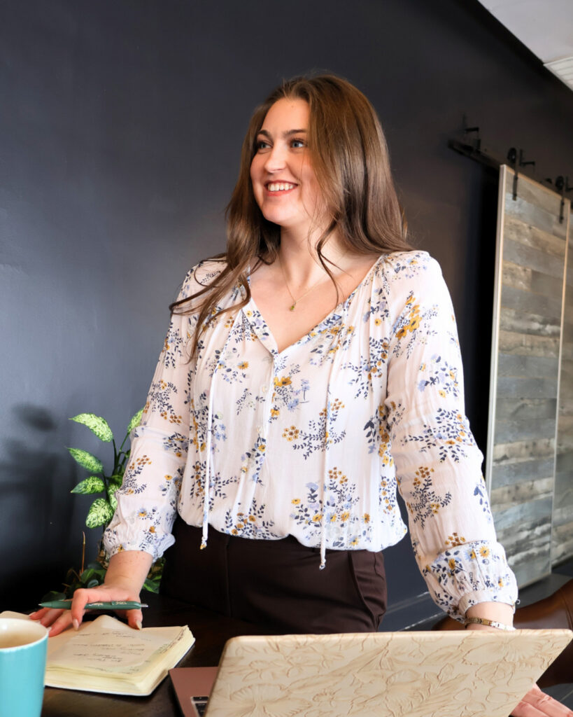 A smiling team member stands at her desk with a notepad and laptop open, ready to assist couples planning their wedding at The Wells Estate.