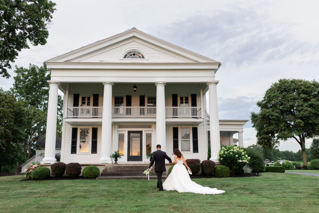 Bride and groom walking hand in hand toward the historic Estate House at The Wells Estate, showcasing classic white columns, manicured lawns, and timeless wedding venue architecture.