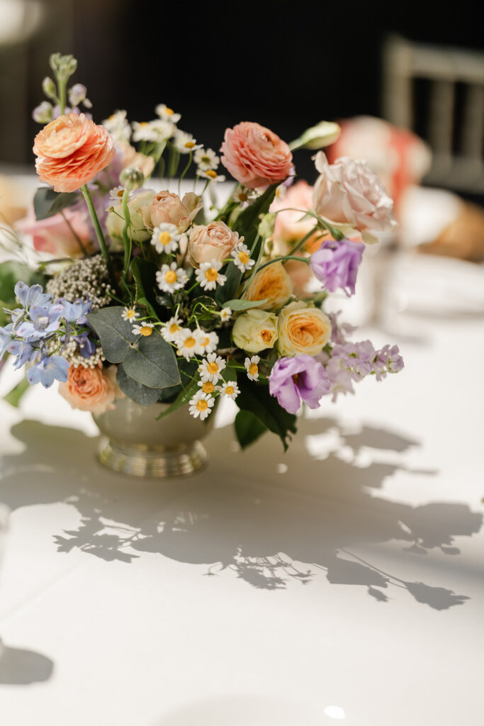 Vibrant garden-style wedding centerpiece featuring ranunculus, roses, chamomile, and soft pastel florals arranged in a low vessel on a sunlit reception table.