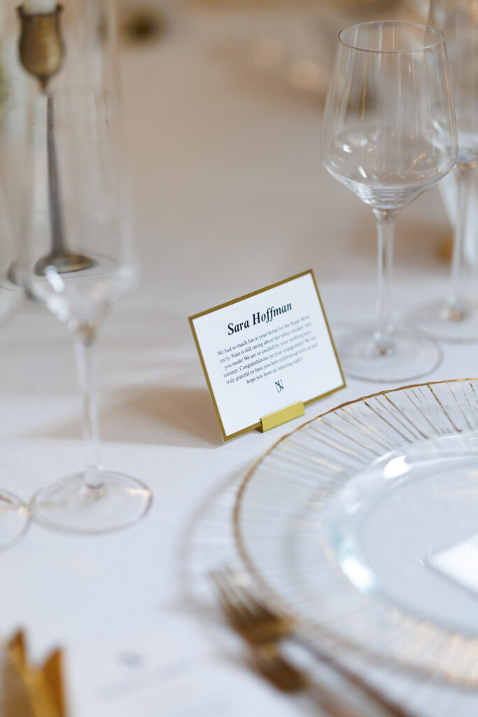 Close-up of elegant wedding table setting featuring a personalized place card, gold-rimmed charger plate, crystal glassware, and soft neutral linens for a refined reception design.