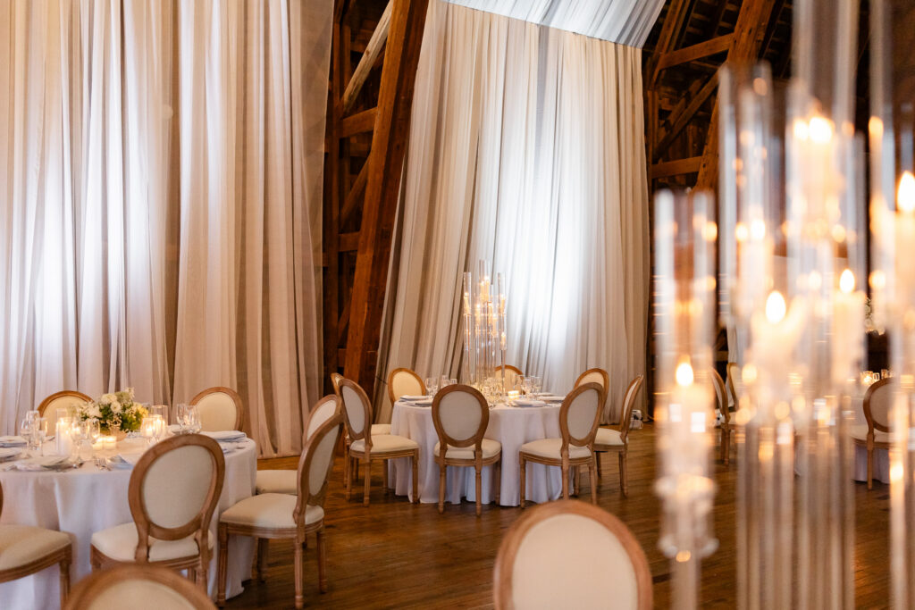 Romantic barn wedding reception at The Wells Estate featuring soft draped ceilings, round tables with neutral linens, candlelight centerpieces, and classic upholstered chairs.
