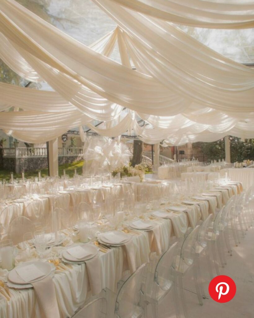 Elegant wedding reception inside a tent with soft draped ceiling fabric, long banquet tables, neutral linens, and clear chairs creating a romantic airy atmosphere.