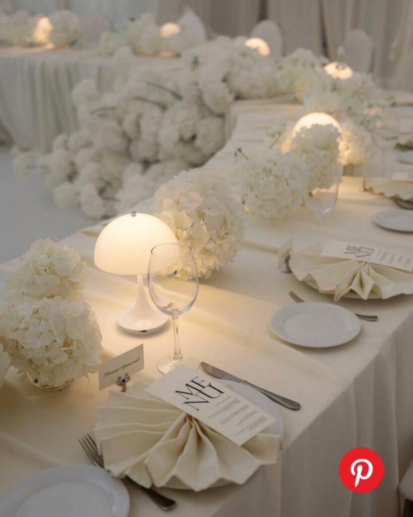 Intimate micro wedding table with white hydrangea arrangements, soft glowing table lamps, folded napkins, and neutral linens for a modern romantic reception design.