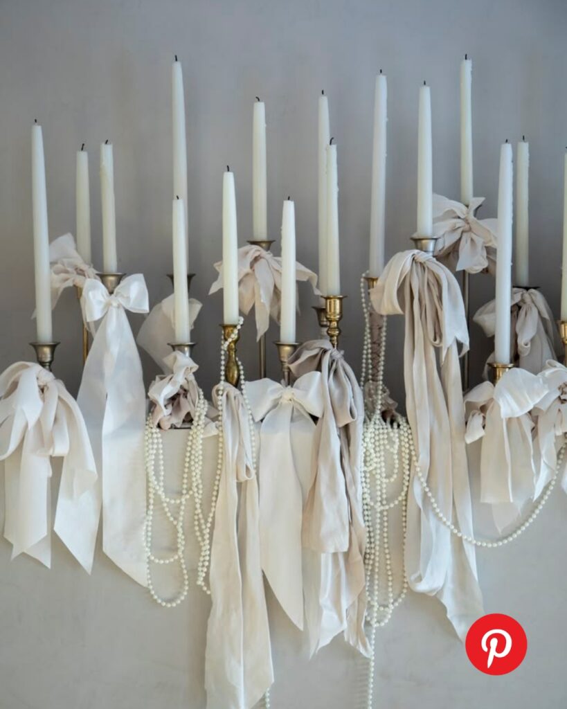 Elegant monochromatic wedding decor featuring tall white taper candles in brass holders adorned with soft silk bows and draped pearl strands.
