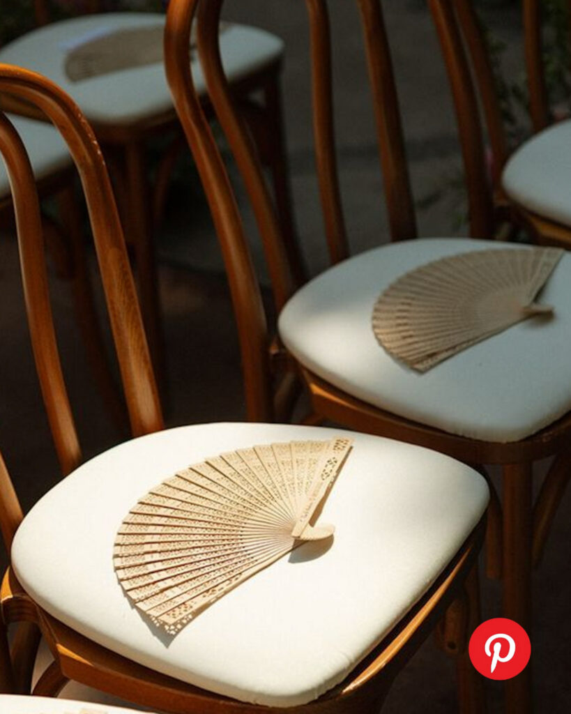 Wooden ceremony chairs with white cushions arranged outdoors, each seat styled with a handheld fan for guest comfort during a summer wedding ceremony.
