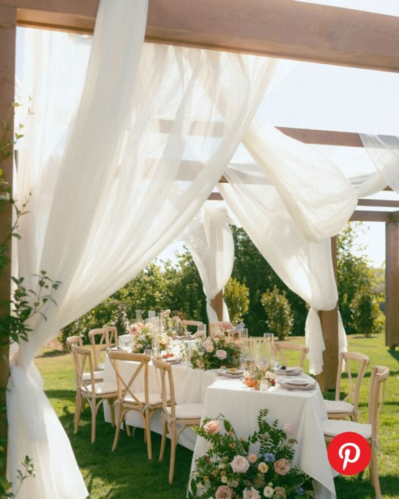 Outdoor wedding reception under a wooden pergola with flowing sheer white drapery, soft garden florals, and an elegant tablescape set on lush green lawn.