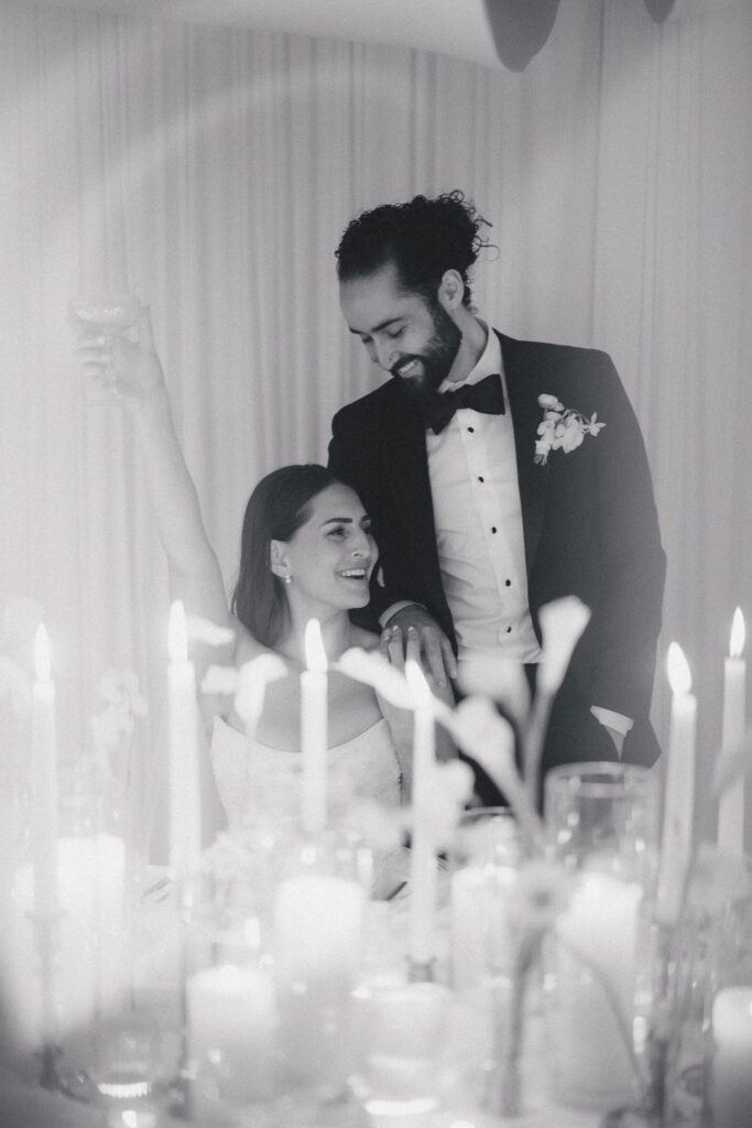 Black and white wedding portrait of a bride and groom smiling together surrounded by candlelit table decor, creating an intimate and timeless reception moment.