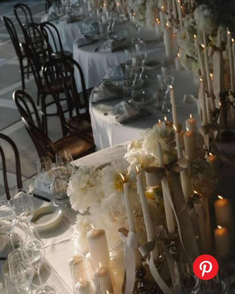 Moody candlelit wedding table with white floral arrangements, layered taper candles, soft linens, and dark wood chairs creating an intimate, romantic reception setting.