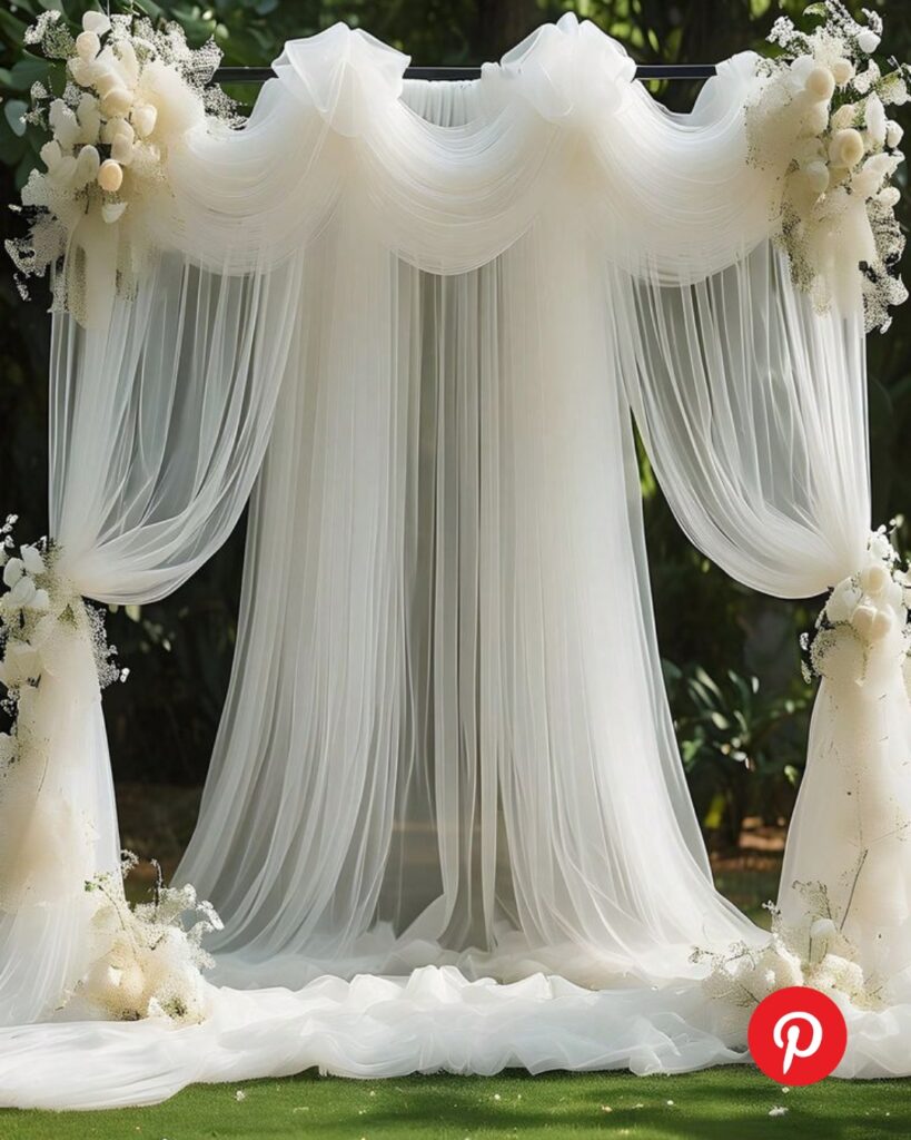 Wedding ceremony arch with layered sheer white drapery and soft floral arrangements, creating a romantic and ethereal backdrop for an outdoor ceremony.