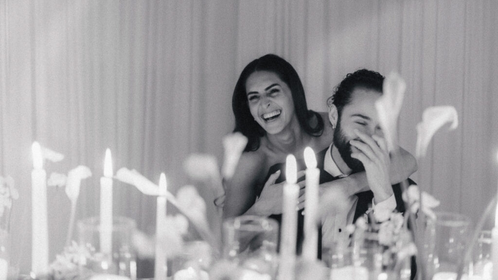 Black and white photo of a couple laughing together at their wedding reception table surrounded by candles and soft romantic decor.