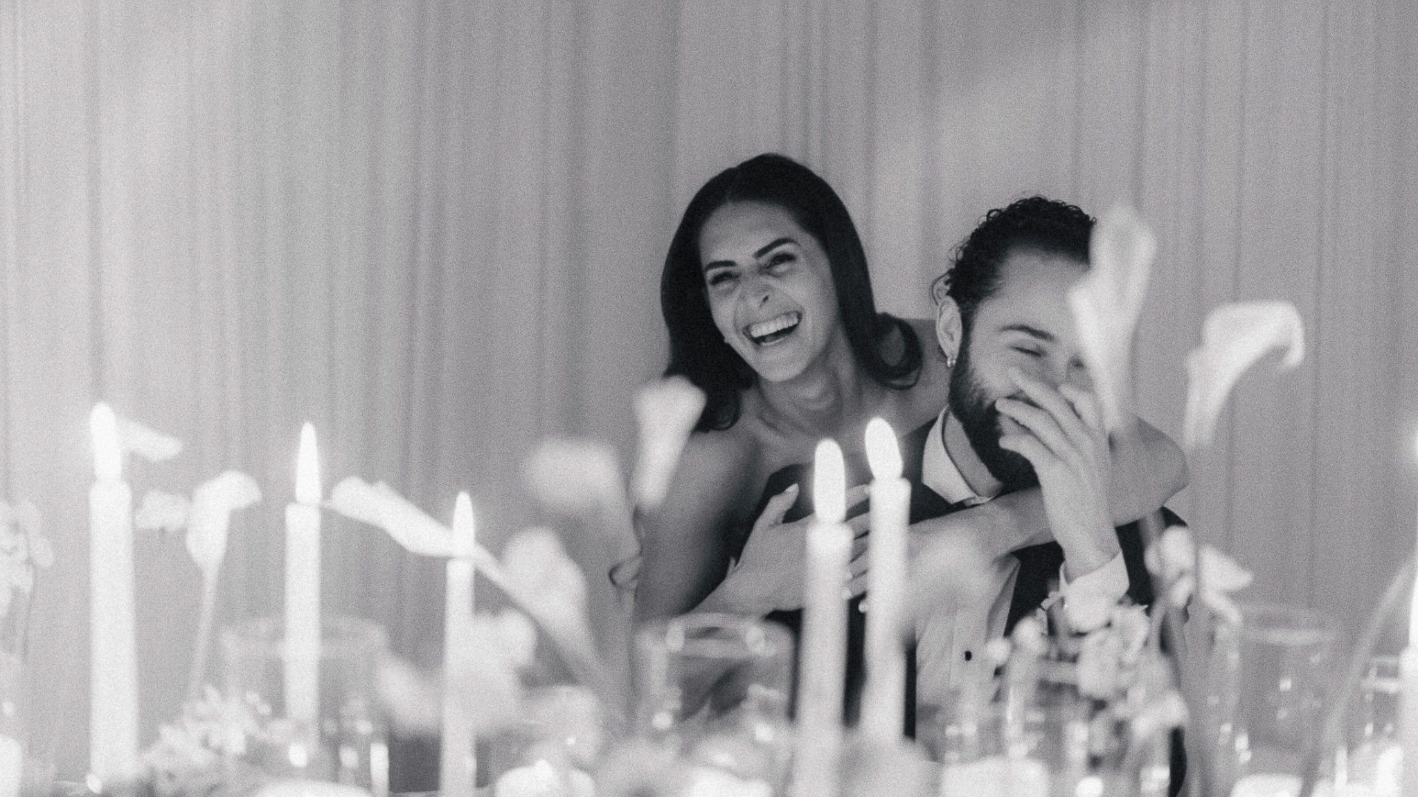 Black and white photo of a couple laughing together at their wedding reception table surrounded by candles and soft romantic decor.
