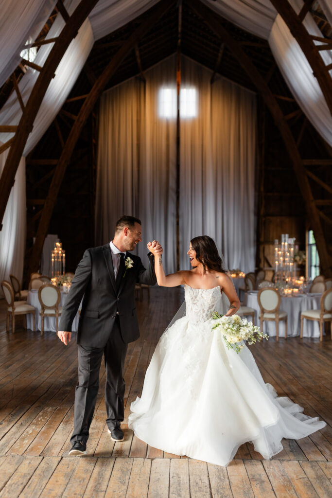 Bride and groom sharing a joyful first dance inside the Countryside Cathedral with wood beams, white draping, and candlelit reception tables at The Wells Estate in Upstate New York.