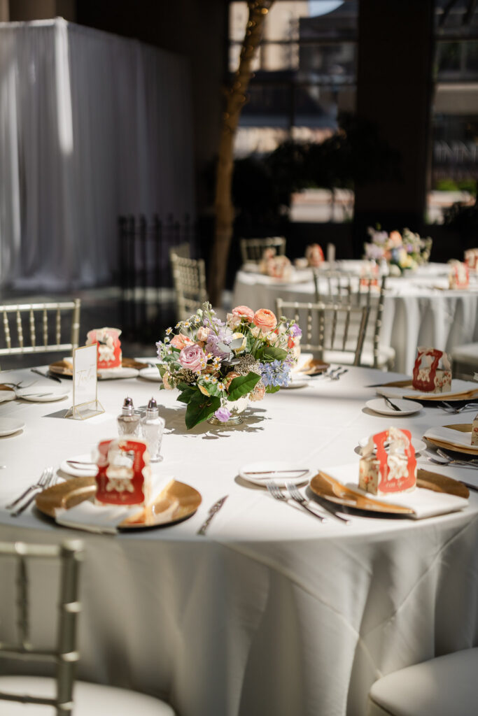 Wedding reception table with white linens, gold charger plates, pastel floral centerpiece, and guest favors, styled in a light-filled indoor venue space.