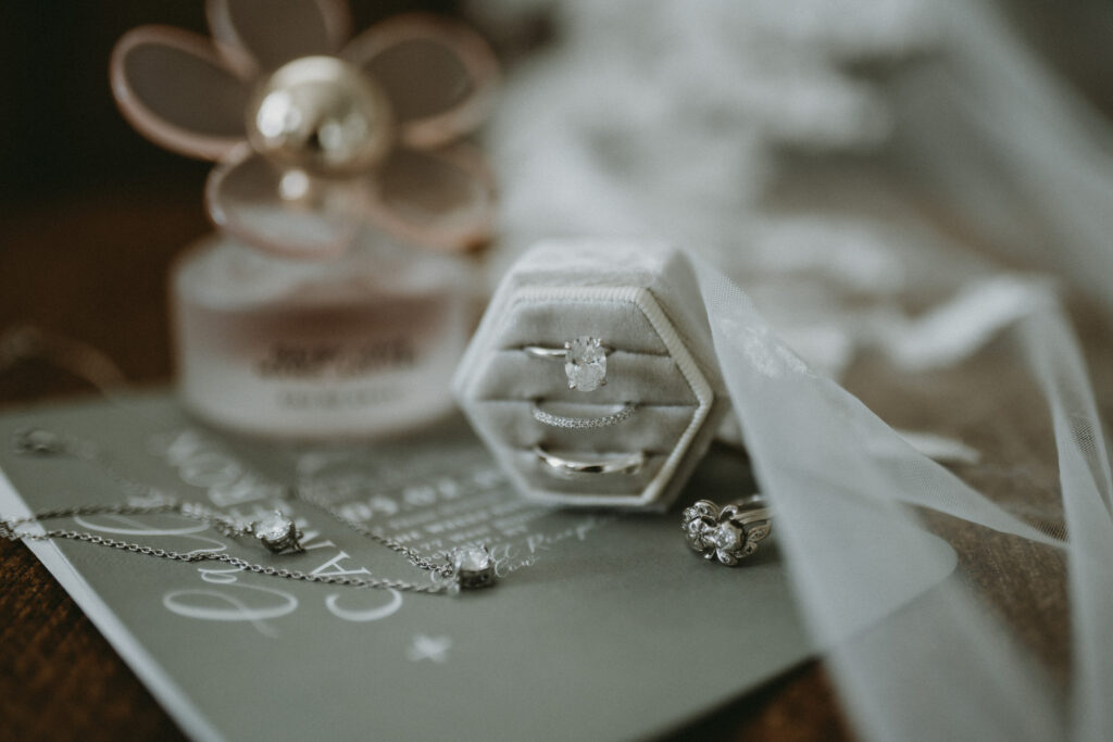 Close-up of wedding rings in a velvet box with invitation suite, perfume, and bridal accessories styled for a romantic wedding detail flat lay.