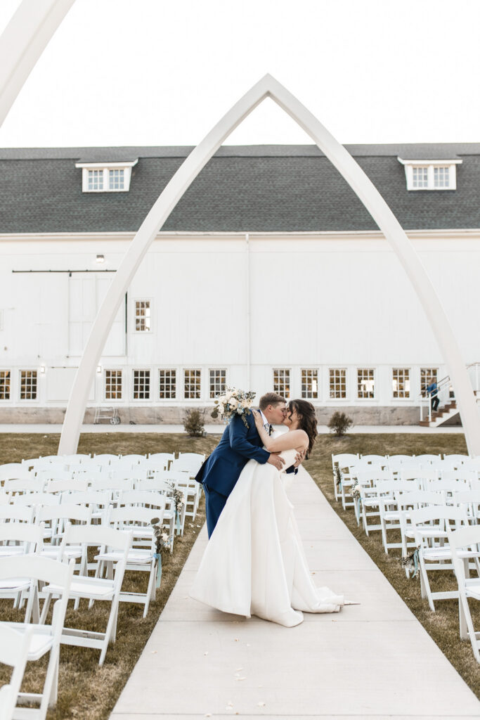 Couple sharing a kiss in the ceremony aisle beneath the Grand Arches with white chairs set for an outdoor wedding.