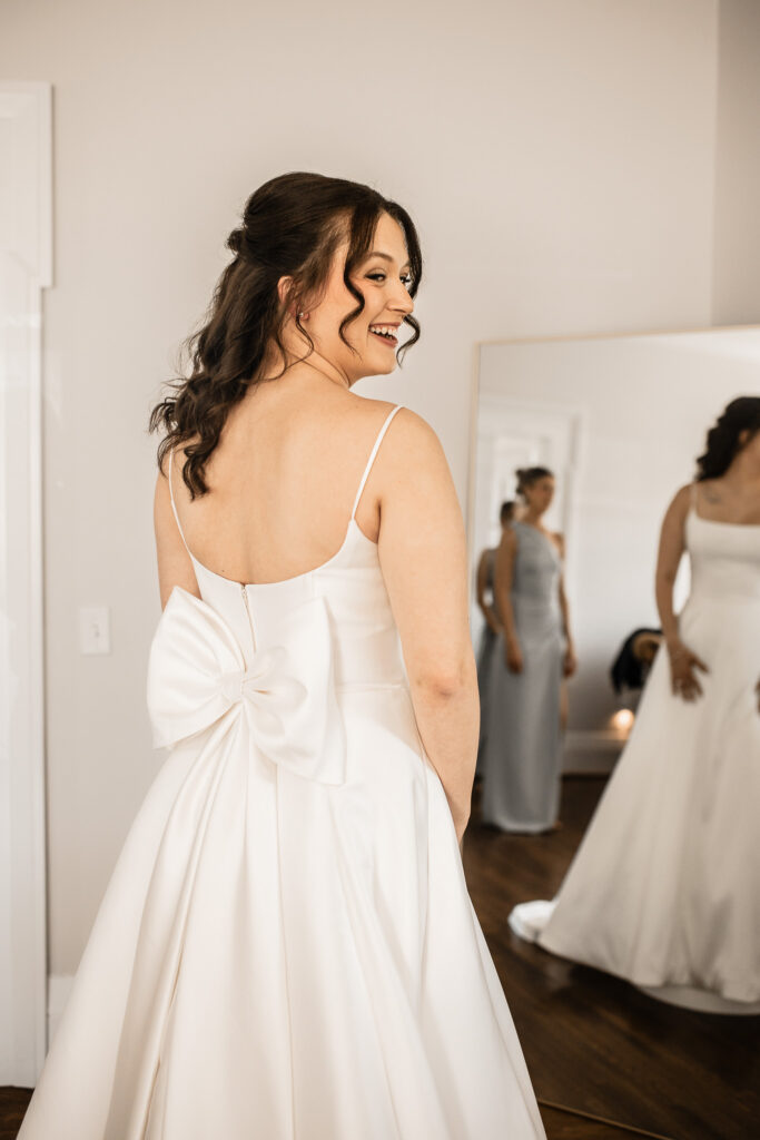 Bride smiling over her shoulder in a wedding dress with a large bow detail on the back inside the bridal suite at The Wells Estate.