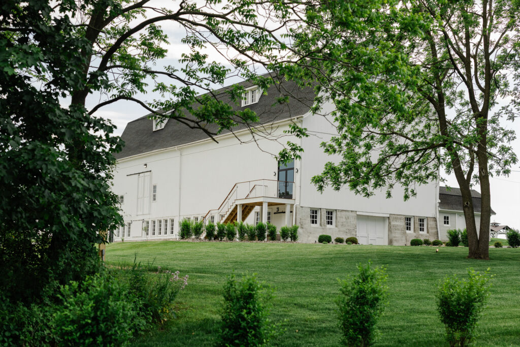 Wide exterior view of The Wells Estate barn with white siding, surrounded by trees and open lawn in a countryside setting.