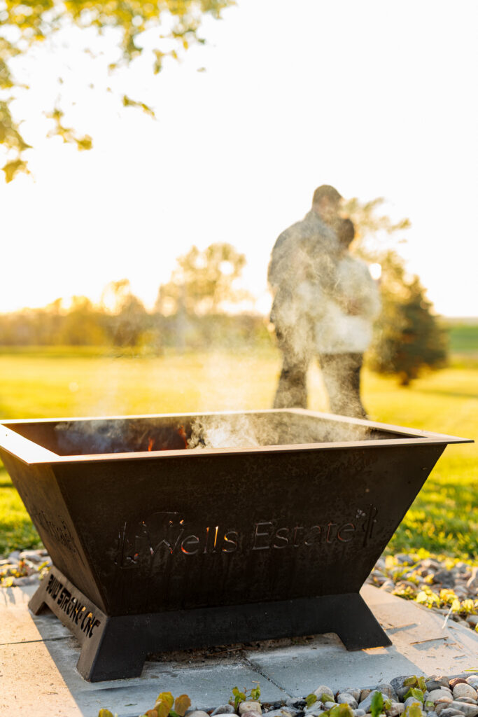 Smoking fire pit with The Wells Estate branding and a couple standing together in the background at sunset.