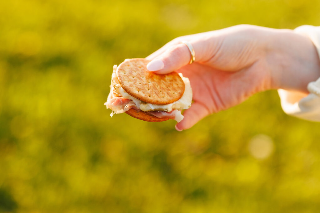 Close-up of a savory s’more style bite with crackers, cheese, and prosciutto held in hand during golden hour.