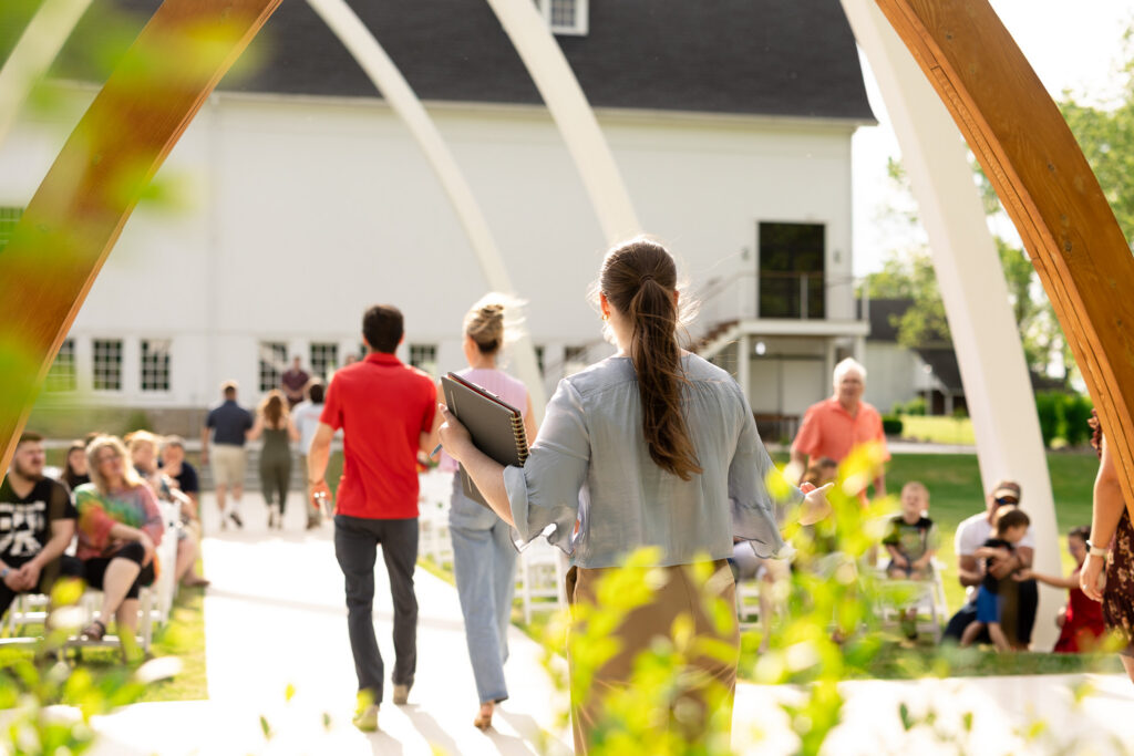 Venue coordinator holding a notebook and guiding guests through a ceremony rehearsal under the Grand Arches at The Wells Estate.