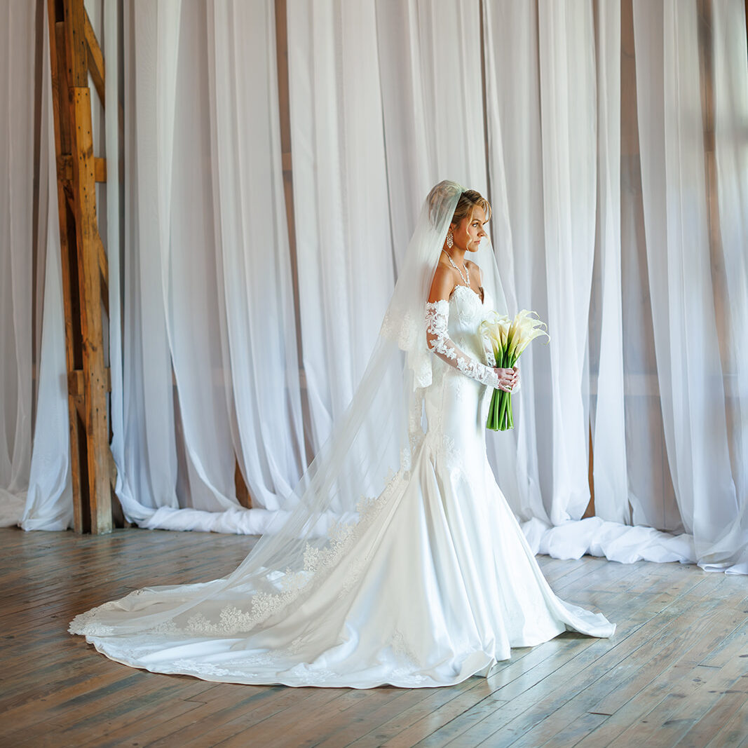 Bride in a lace wedding gown and cathedral-length veil holding a bouquet of white calla lilies inside the Countryside Cathedral at The Wells Estate, surrounded by floor-to-ceiling white draping and rustic wood details.