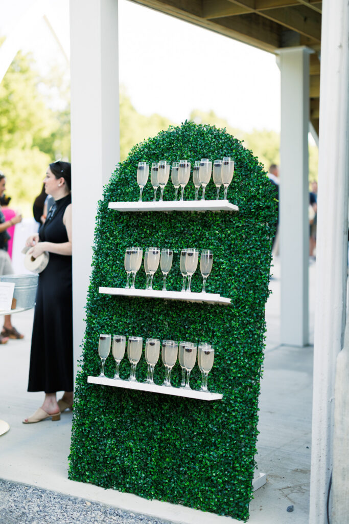 A champagne welcome wall with sparkling wine displayed on greenery-covered shelves during cocktail hour at The Wells Estate wedding venue in Avon, New York.
