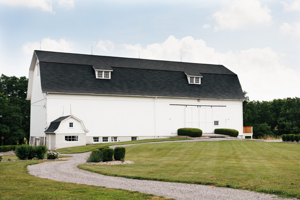 Wide exterior view of The Wells Estate's iconic white wedding barn surrounded by manicured grounds and a winding gravel drive in Avon, New York.