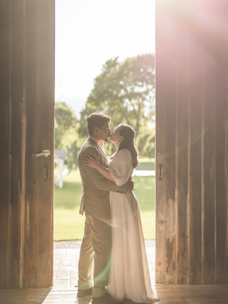 Couple shares a kiss framed by open barn doors with warm sunlight pouring into The Wells Estate reception space.