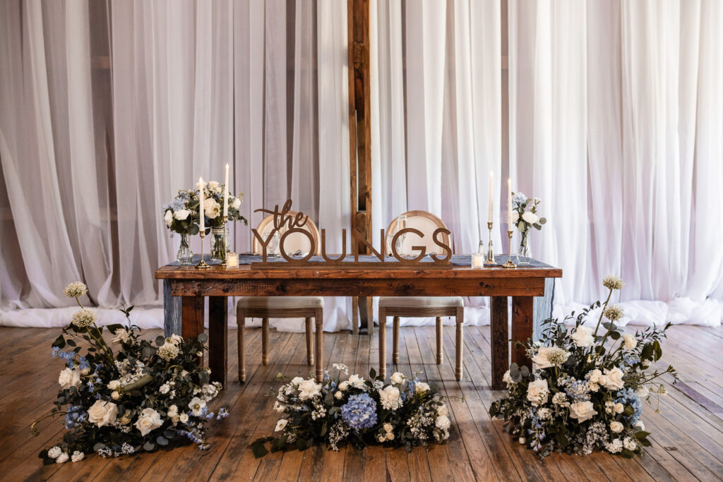 Reception sweetheart table setup featuring floral arrangements, candles, and “The Youngs” signage inside the barn at The Wells Estate.