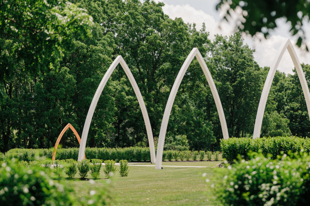 The iconic Grand Arches ceremony site at The Wells Estate surrounded by manicured landscaping and mature trees, showcasing the venue's signature outdoor wedding ceremony space in Avon, New York.