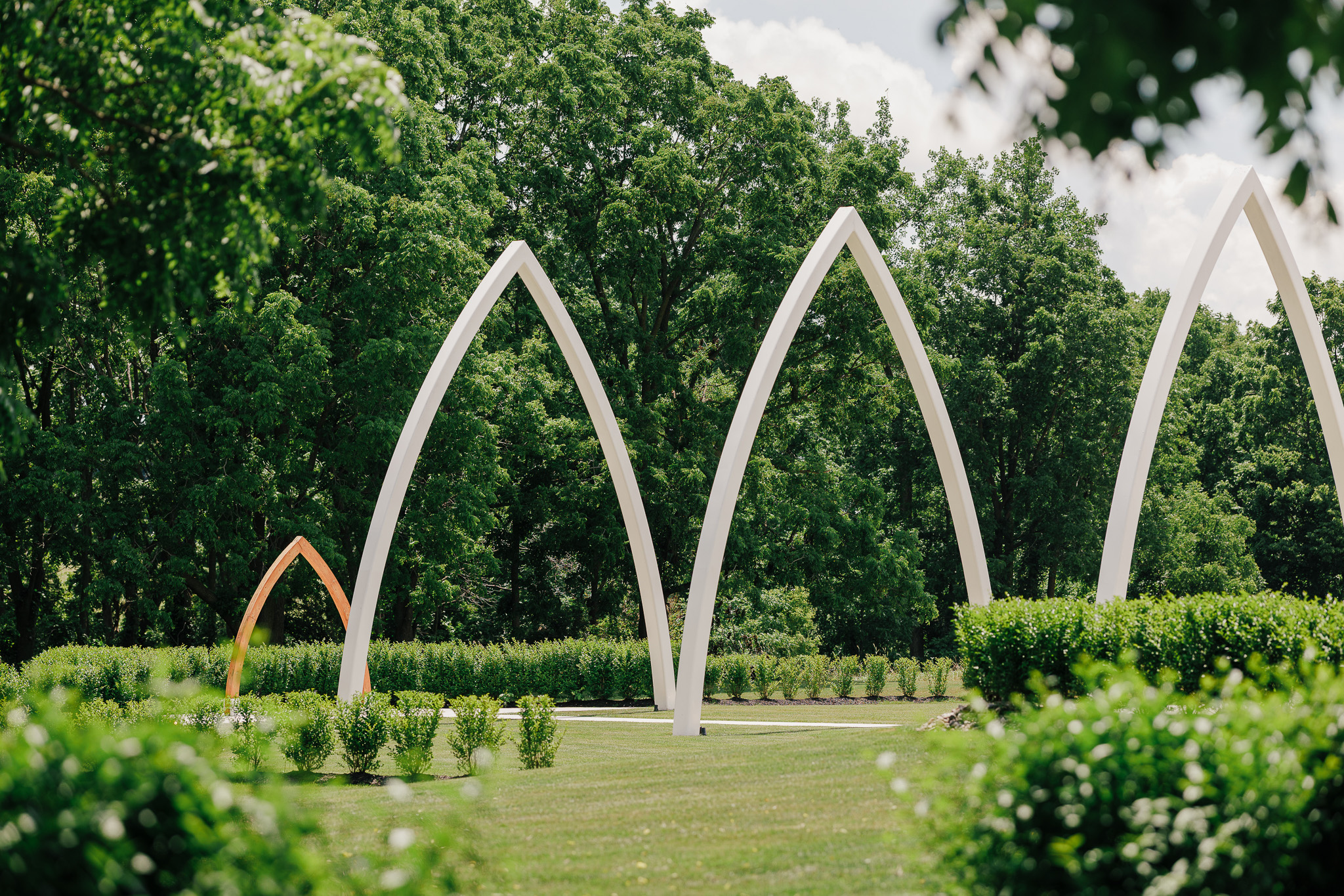 The iconic Grand Arches ceremony site at The Wells Estate surrounded by manicured landscaping and mature trees, showcasing the venue's signature outdoor wedding ceremony space in Avon, New York.