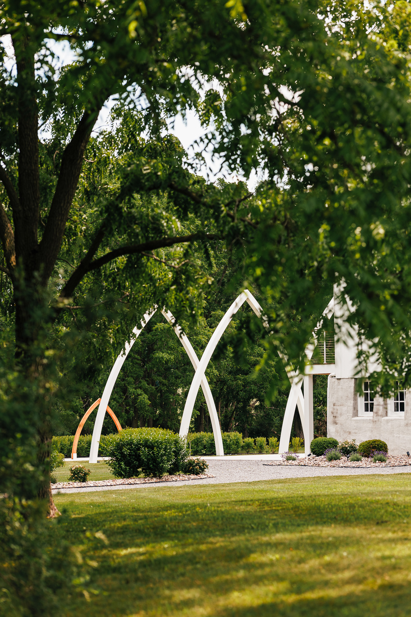 The signature Grand Arches ceremony site at The Wells Estate viewed through mature trees, showcasing the modern white arches surrounded by lush landscaping.