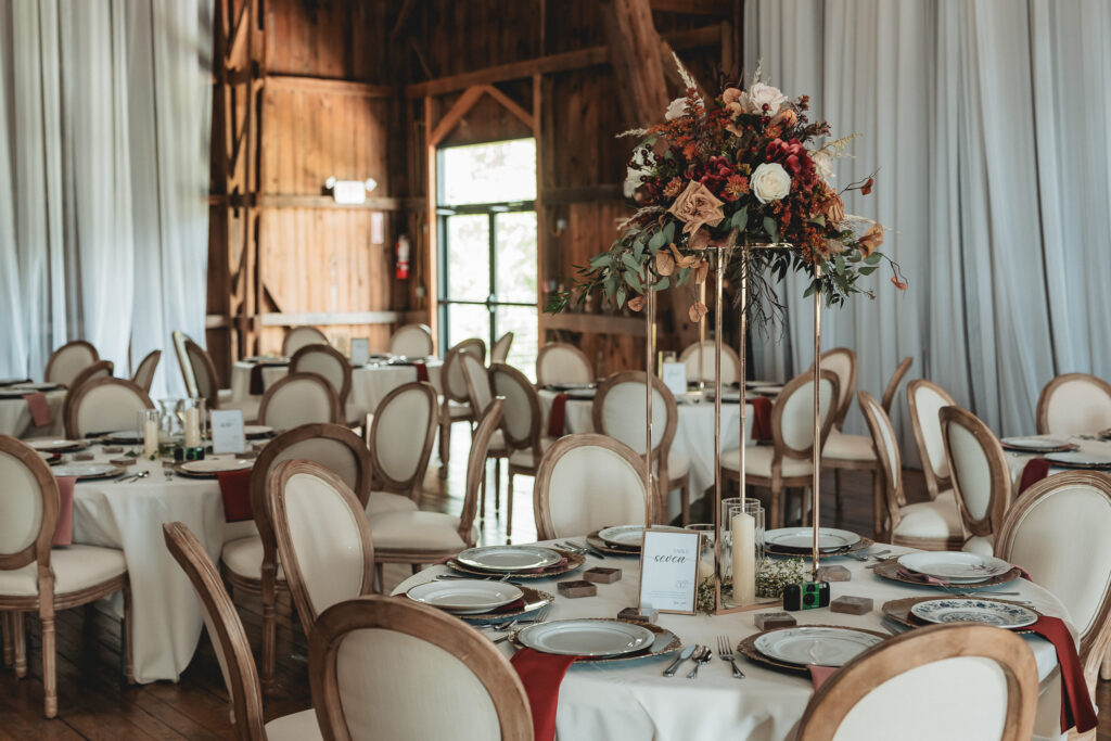 Elegant wedding reception setup inside The Wells Estate barn featuring round tables, neutral linens, wood chairs, and tall fall-toned floral centerpieces.