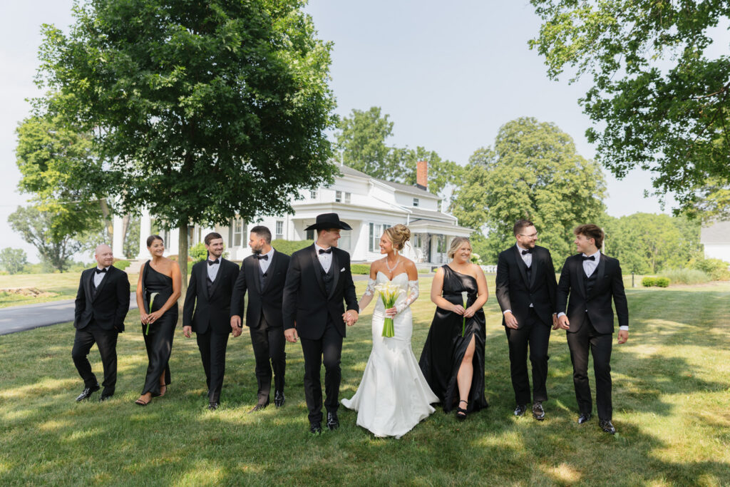 Bride and groom walking with their wedding party in black formal attire across the lawn with the Estate House at The Wells Estate in the background.