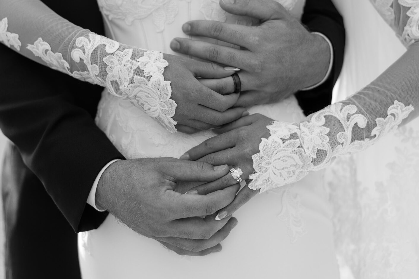 Black-and-white detail portrait of a bride and groom embracing, highlighting the bride’s lace sleeves, engagement ring, wedding band, and the groom’s wedding ring at The Wells Estate.