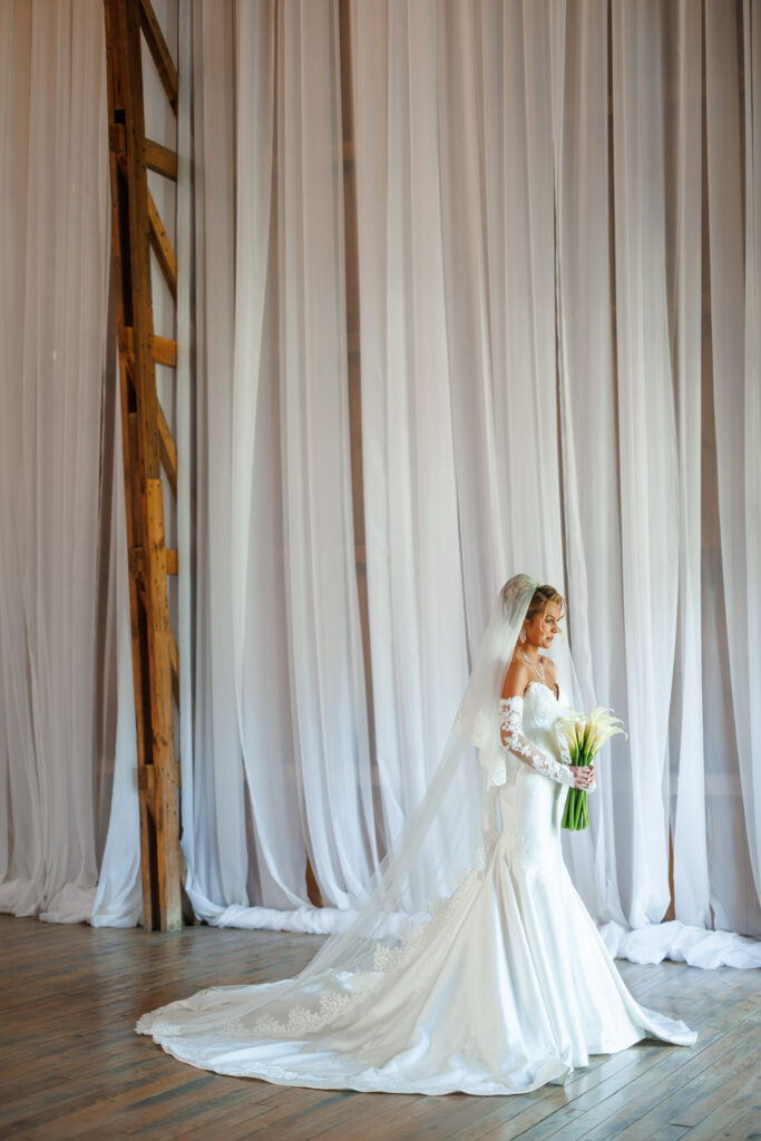 Bride holding a white calla lily bouquet in front of floor-to-ceiling white draping inside the Countryside Cathedral at The Wells Estate.