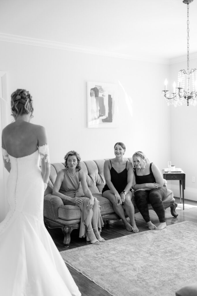 Black-and-white photo of the bride revealing her wedding dress to three women seated together during wedding preparations at The Wells Estate.