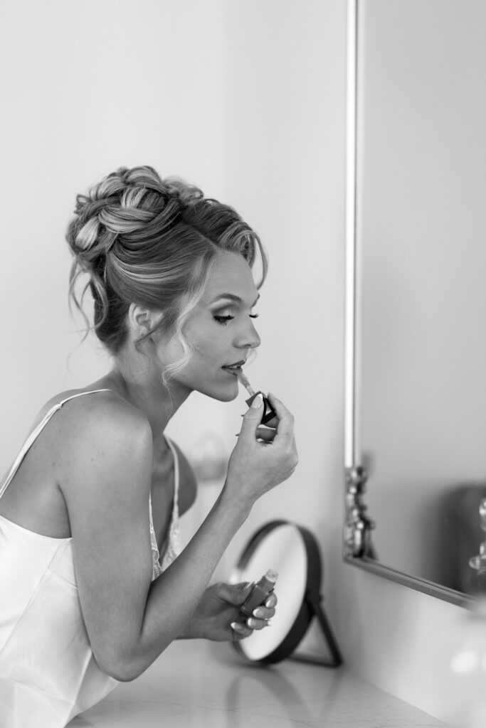 Black-and-white portrait of the bride applying lip color at a mirror while getting ready for her wedding at The Wells Estate.