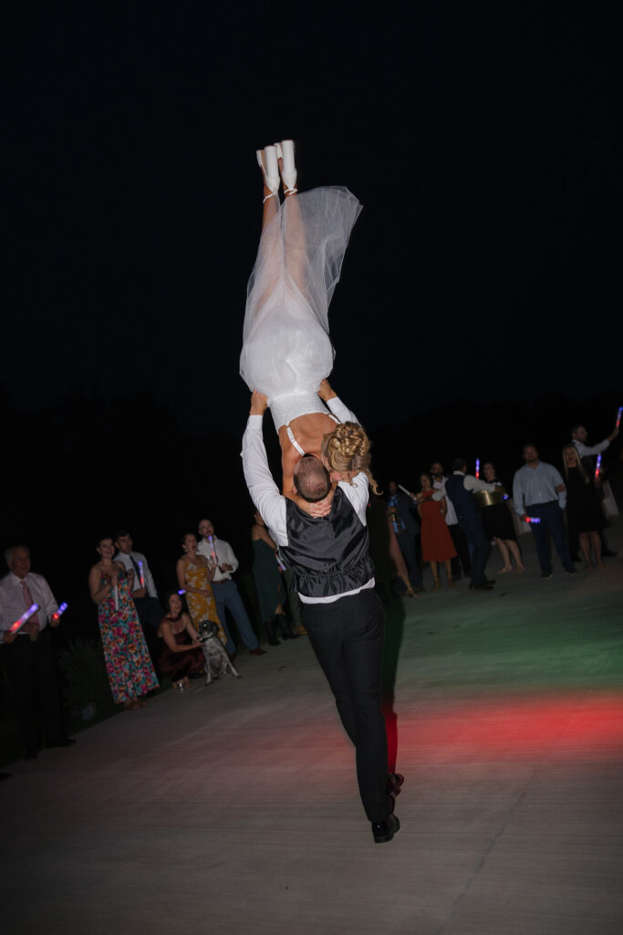 Groom lifts the bride into the air during a lively nighttime wedding reception on the outdoor dance floor at The Wells Estate as guests cheer around them.