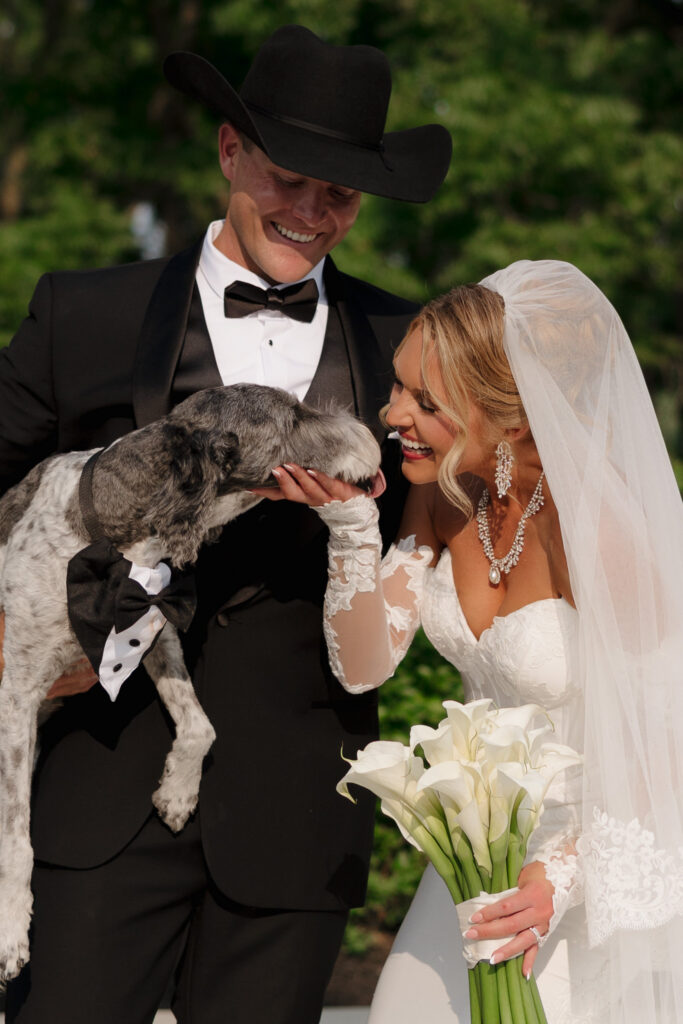 Bride holding a white calla lily bouquet as she and the groom pose with their dog dressed in a tuxedo at The Wells Estate.