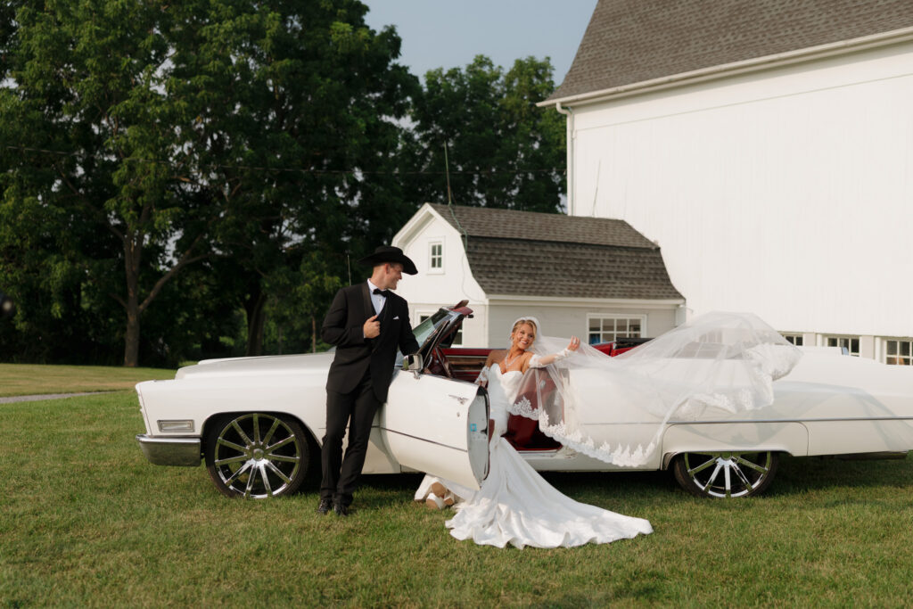 Bride and groom posing beside a white vintage convertible on the lawn near the Countryside Cathedral at The Wells Estate.