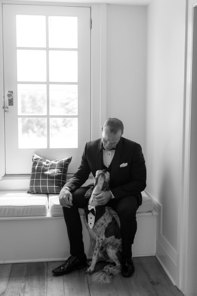 Black-and-white photo of the groom in a tuxedo sitting with his dog dressed in a bow tie during wedding preparations at The Wells Estate.