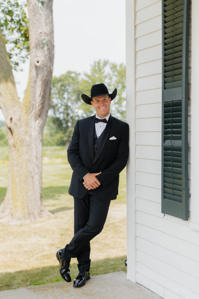 Groom in a black tuxedo and cowboy hat posing beside the white exterior and dark shutters of The Wells Estate.