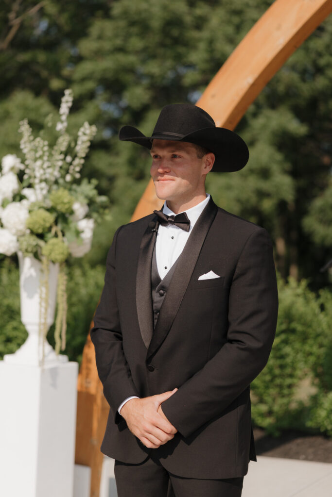 Groom in a black tuxedo and cowboy hat waiting beneath the Grand Arches for an outdoor wedding ceremony at The Wells Estate.