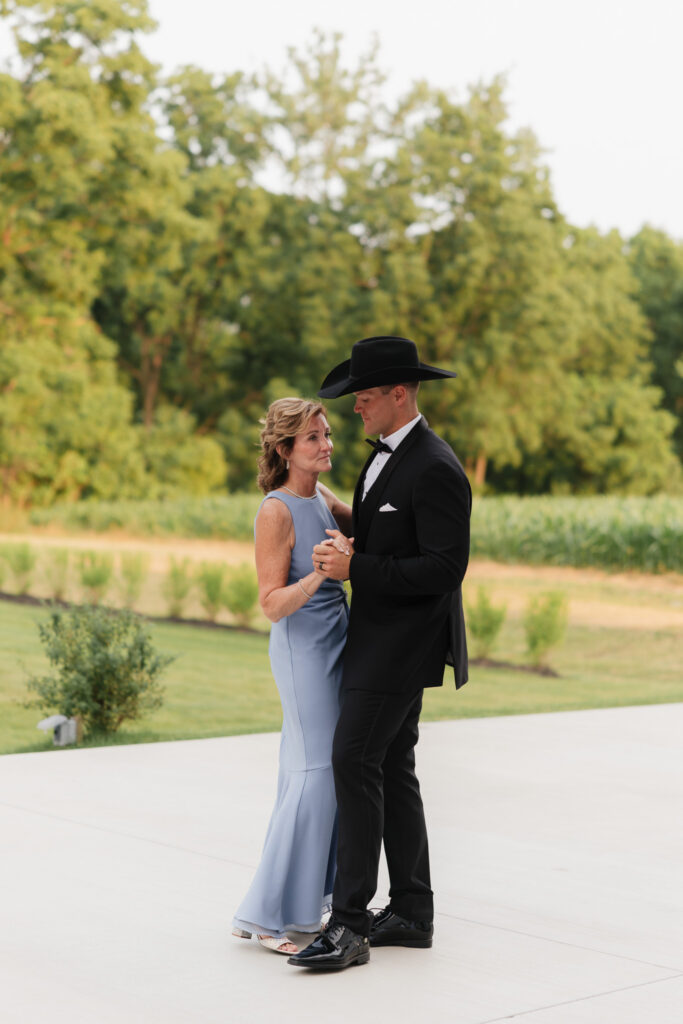Groom in a black tuxedo and cowboy hat shares a mother-son dance during an outdoor wedding reception at The Wells Estate.