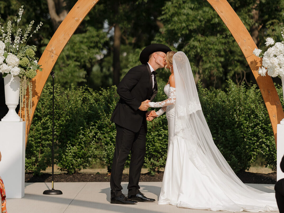 Bride and groom share their first kiss beneath a wooden pointed arch decorated with white florals during an outdoor wedding ceremony at The Wells Estate.